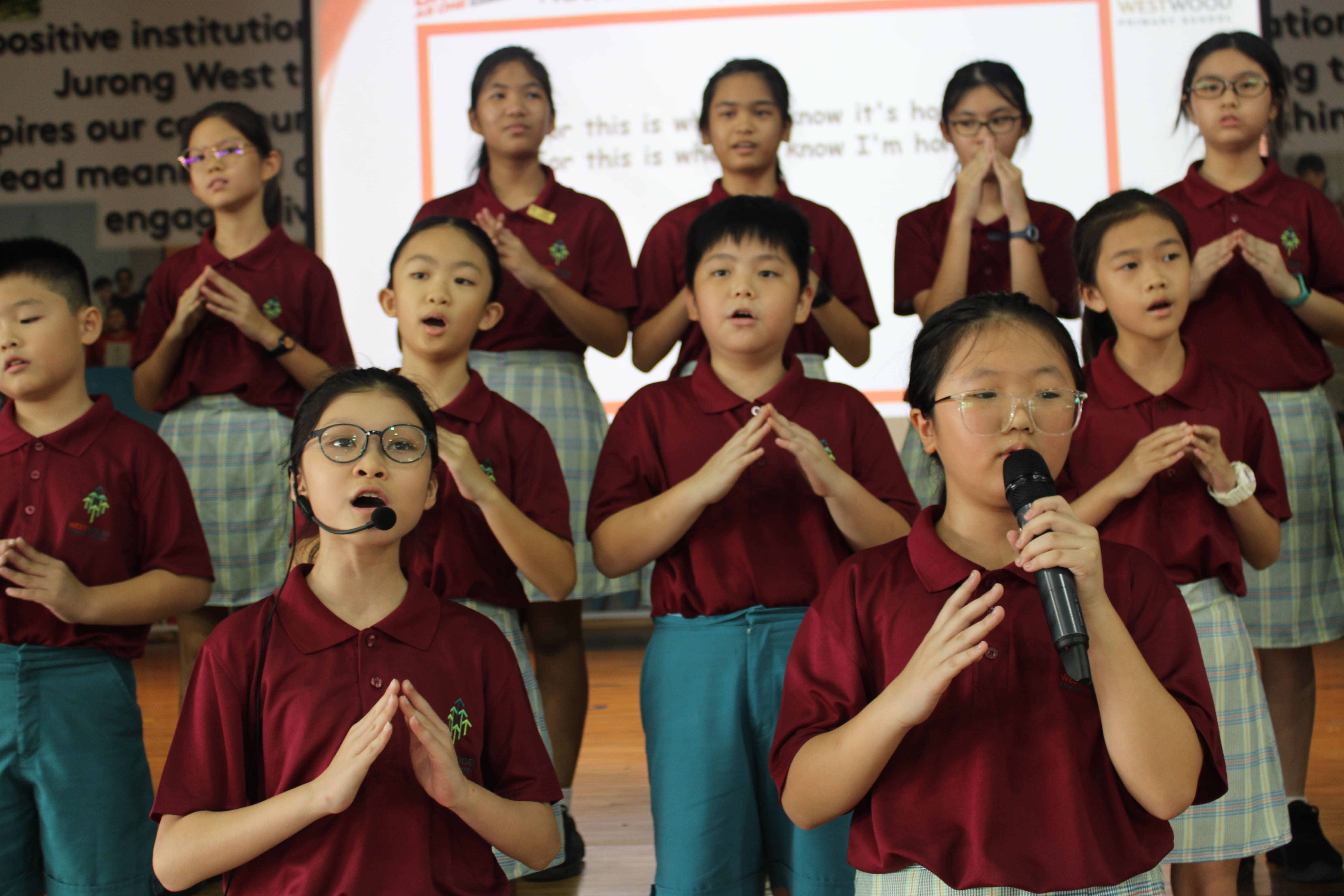 Children in maroon school uniforms perform a choir song on stage, two in front singing into microphones.